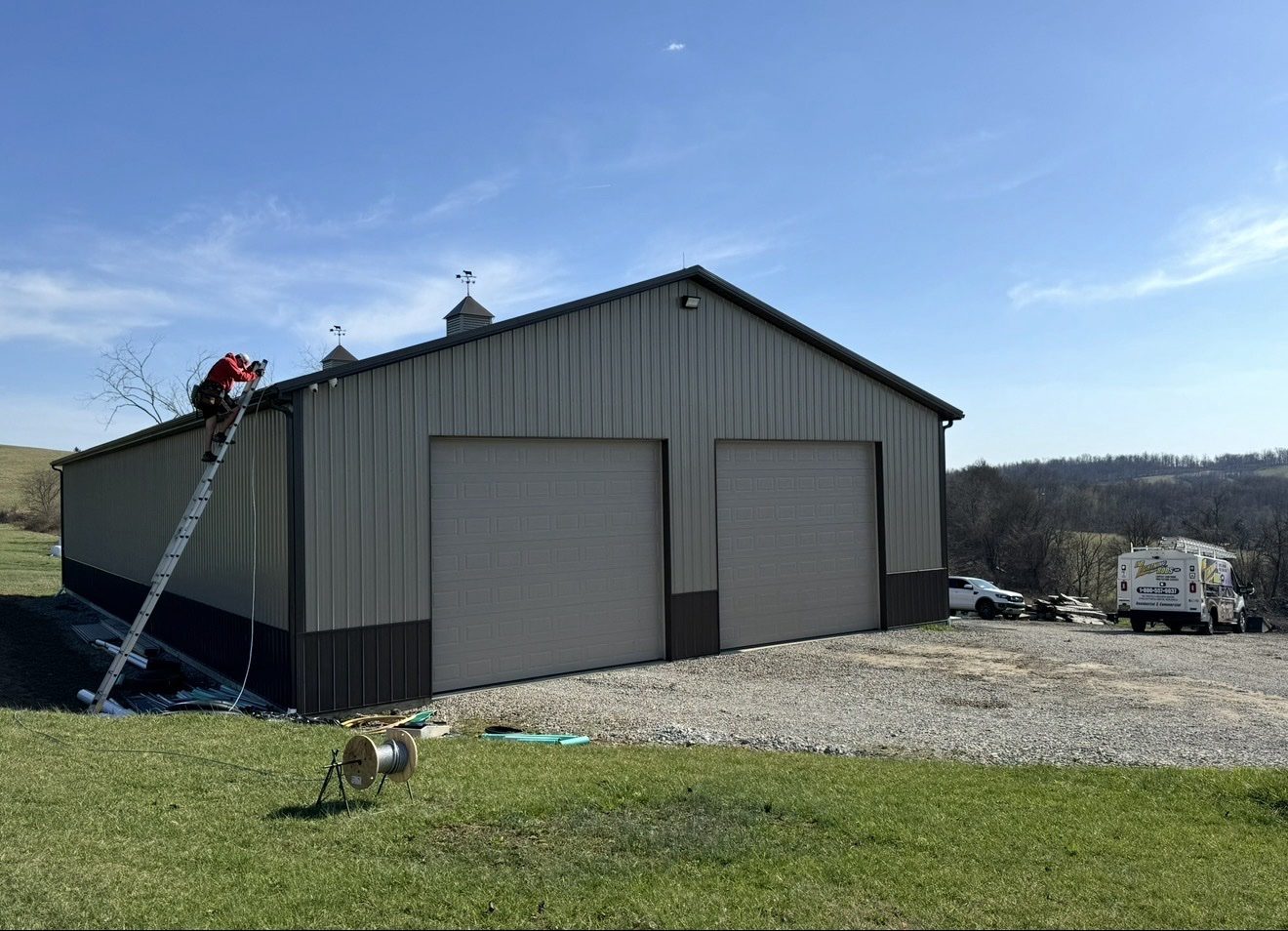 Crew member on a ladder installing lightning protection on a large metal farm shop barn, with the WB Lightning Rods truck parked alongside